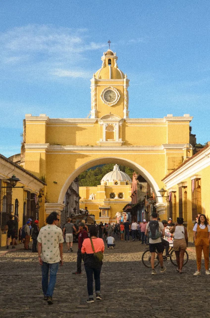 The yellow Santa Catalina Arch on a cobbled street in Antigua, Guatemala, with people walking beneath it.
