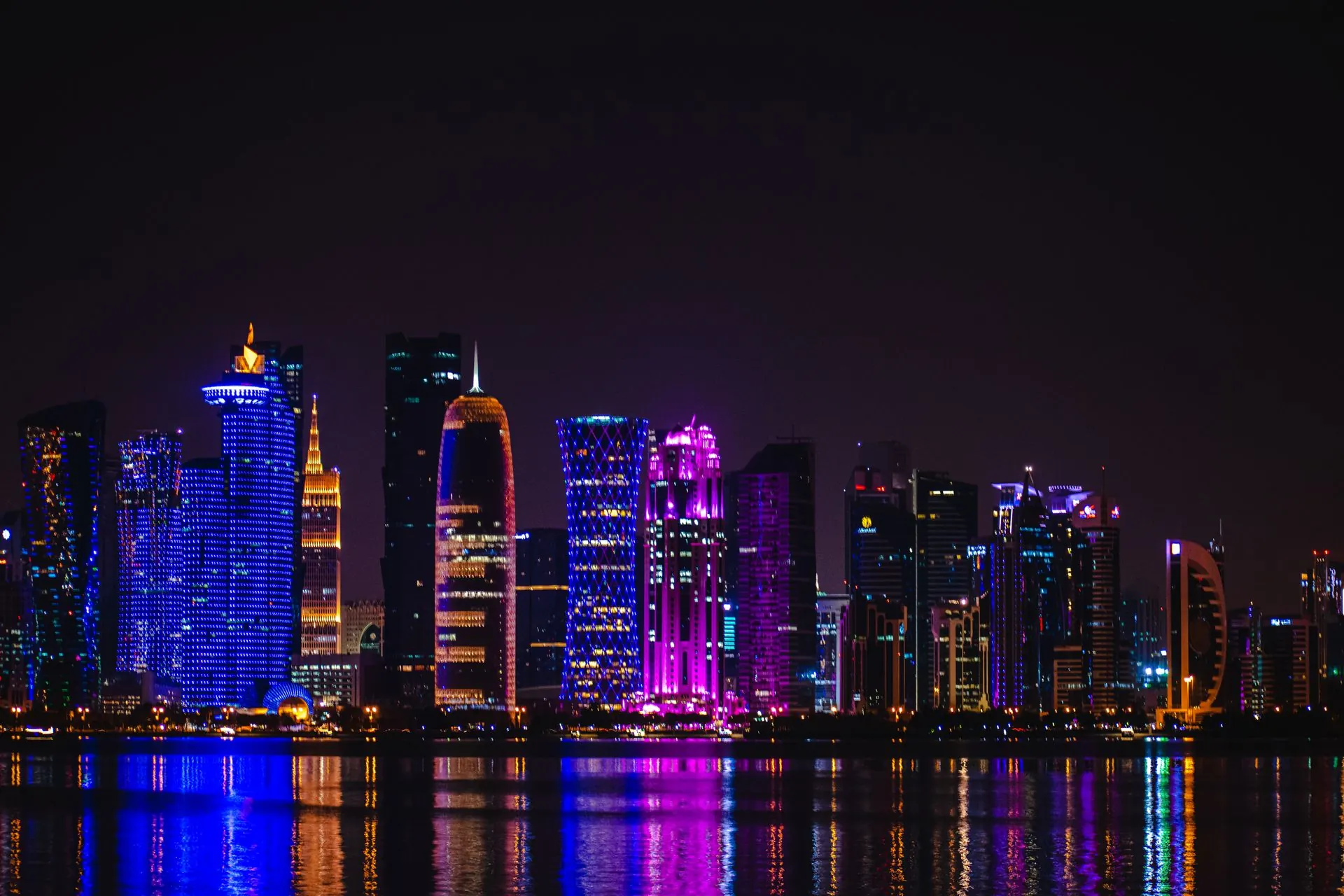 Doha's West Bay towers lit in blue and purple at night, reflected in the water of the bay.